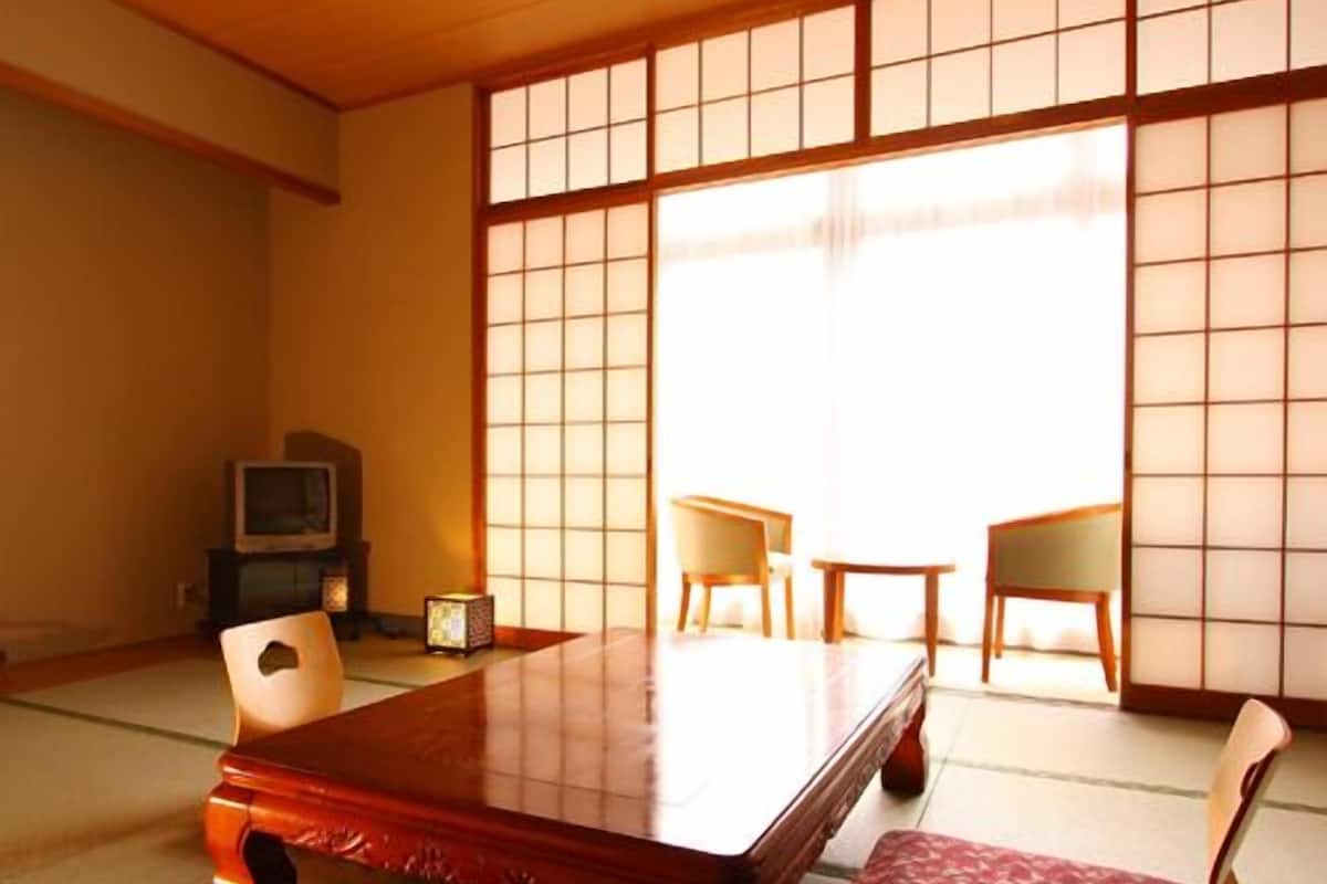 Una habitación japonesa tradicional con suelo de tatami, una mesa de madera baja, dos sillas y un televisor.