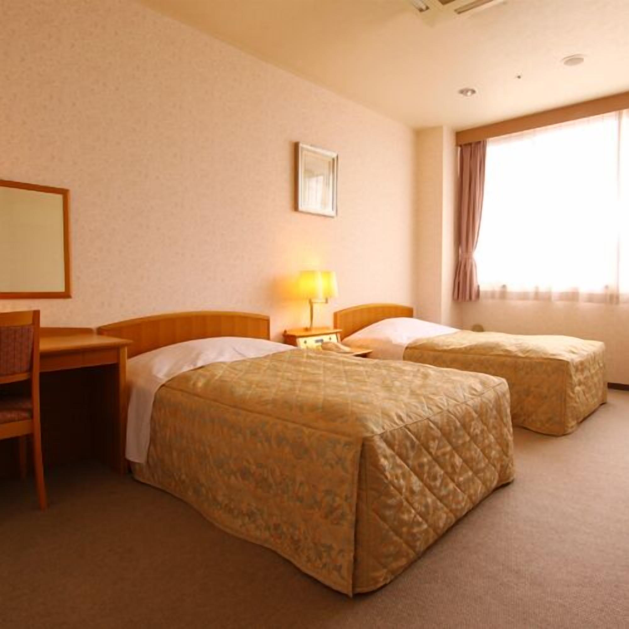Quarto de hotel com duas camas, uma escrivaninha, uma cadeira, um abajur e janela com cortinas.