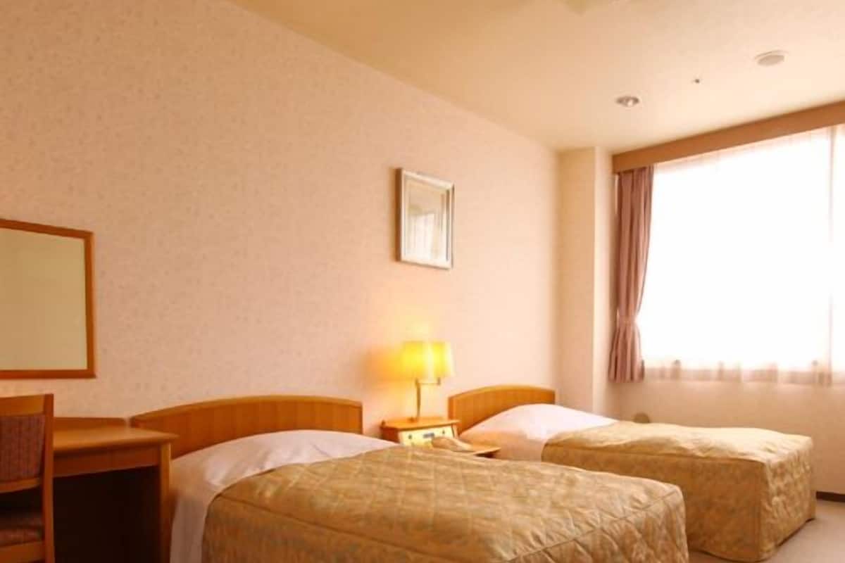 Quarto de hotel com duas camas, uma escrivaninha, uma cadeira, um abajur e janela com cortinas.