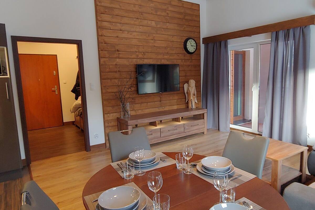 Ein modernes Wohnzimmer mit Holzwand, einem Flachbildfernseher, einem Esstisch für vier Personen und einer gemütlichen Sitzecke mit Sofa und Sesseln.