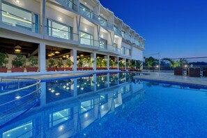 Um hotel com uma grande piscina ao ar livre, várias varandas e áreas de estar internas com plantas em vasos.