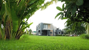 A modern two-story house with a flat roof and a balcony, surrounded by lush green grass and tropical plants. - Malhaar Resort (Dahegam)