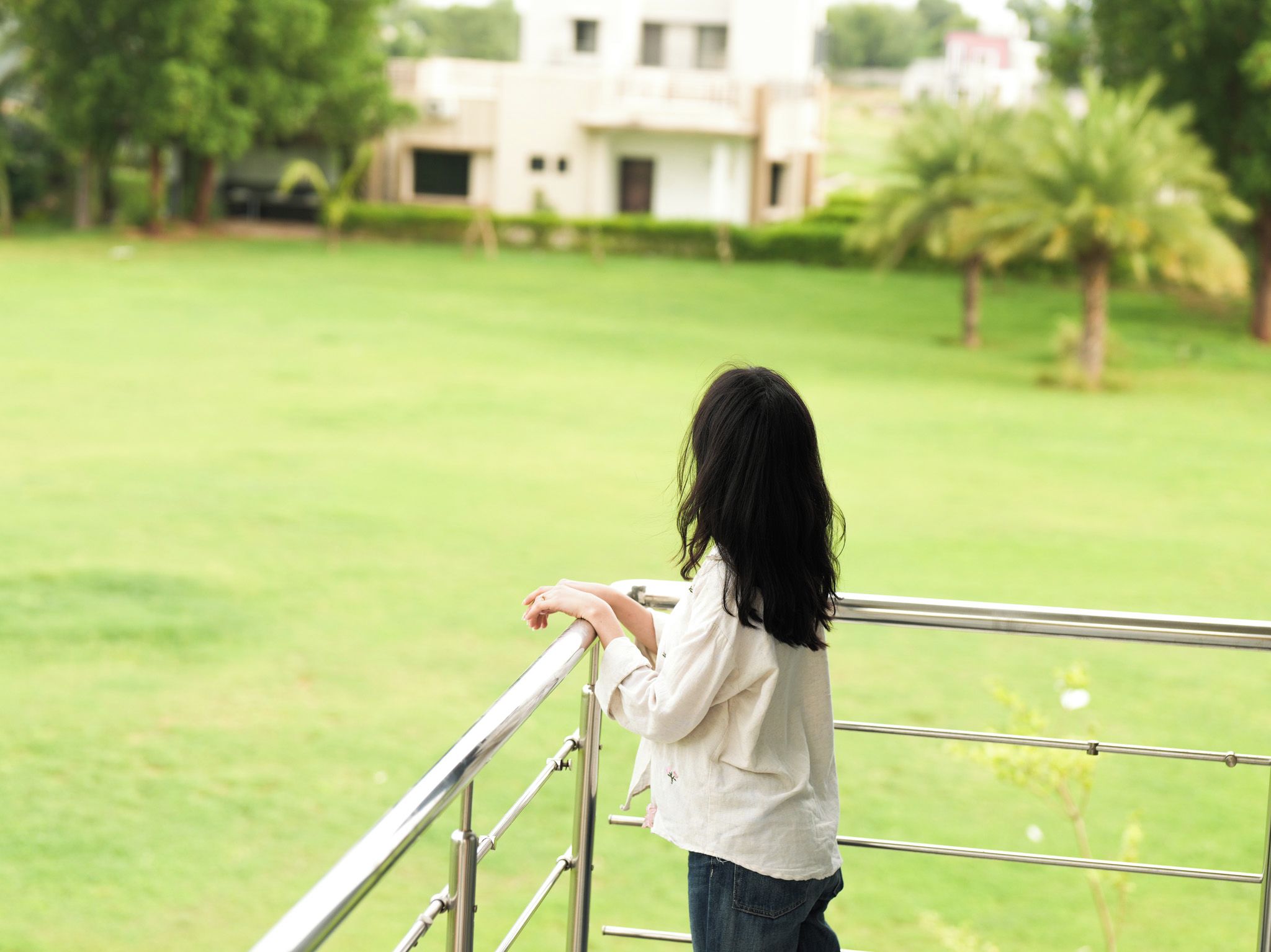 A person standing on a balcony with a view of a green lawn and a building in the background.