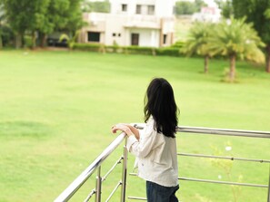 A person standing on a balcony with a view of a green lawn and a building in the background. - Malhaar Resort (Dahegam)