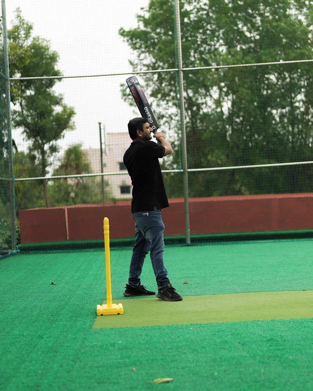 A person practicing cricket on a green artificial turf field with a netted fence in the background.