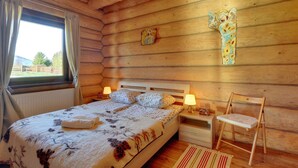 Un dormitorio de cabaña de madera con una cama, dos lámparas de noche, una silla de madera y una cruz en la pared.