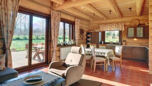 Interior acogedor de una cabaña con comedor, cocina y grandes ventanales que ofrecen vistas del exterior.