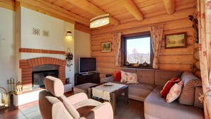 Interior acogedor de una cabaña de troncos con chimenea, un sofá, un televisor y una ventana con vistas a las montañas.