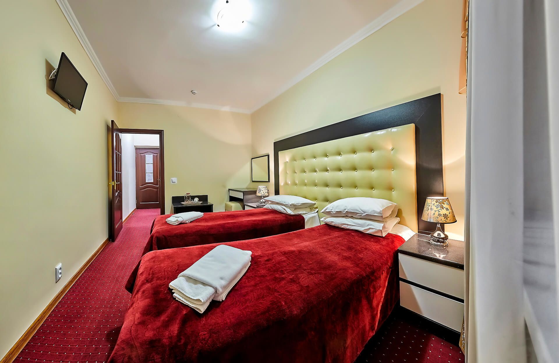 A hotel room with two beds, a large headboard, a bedside table with a lamp, and a red carpeted floor.