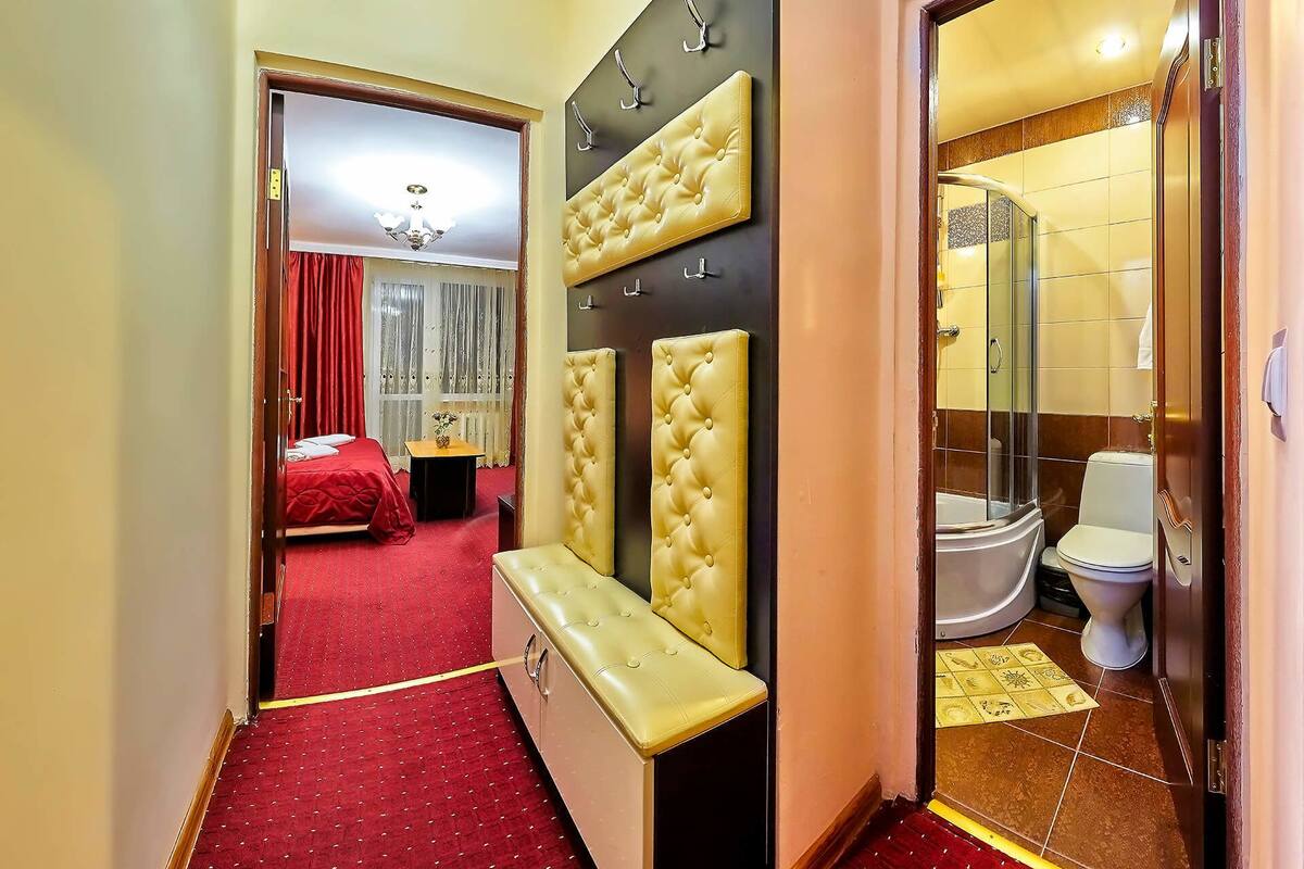 Quarto de hotel com carpete vermelho, cama com roupa de cama vermelha, um banco, televisão presa à parede e banheiro com vaso sanitário e chuveiro.