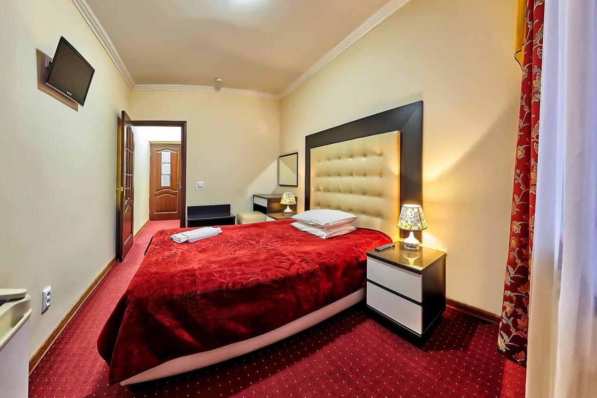 Quarto de hotel com cama grande, mesa de cabeceira, televisão fixada na parede e porta que dá para outro cômodo.