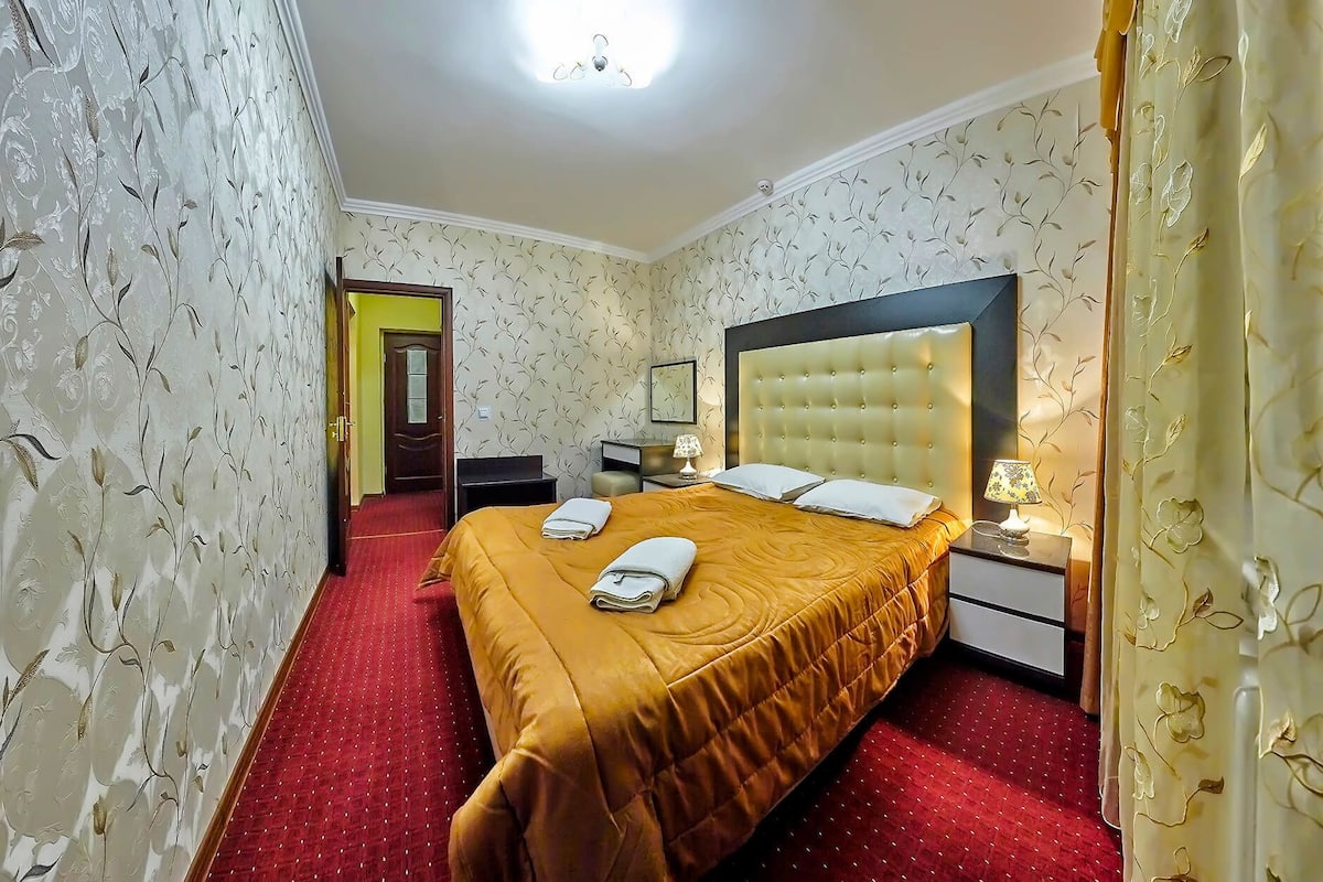 Quarto de hotel com uma cama grande, uma mesa de cabeceira, uma luminária e uma porta que dá para outro cômodo.