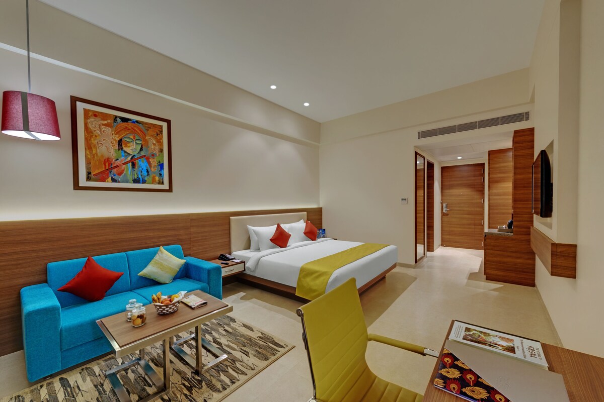 A hotel room with a bed, a sofa, a coffee table, a chair, a TV, and a painting on the wall.