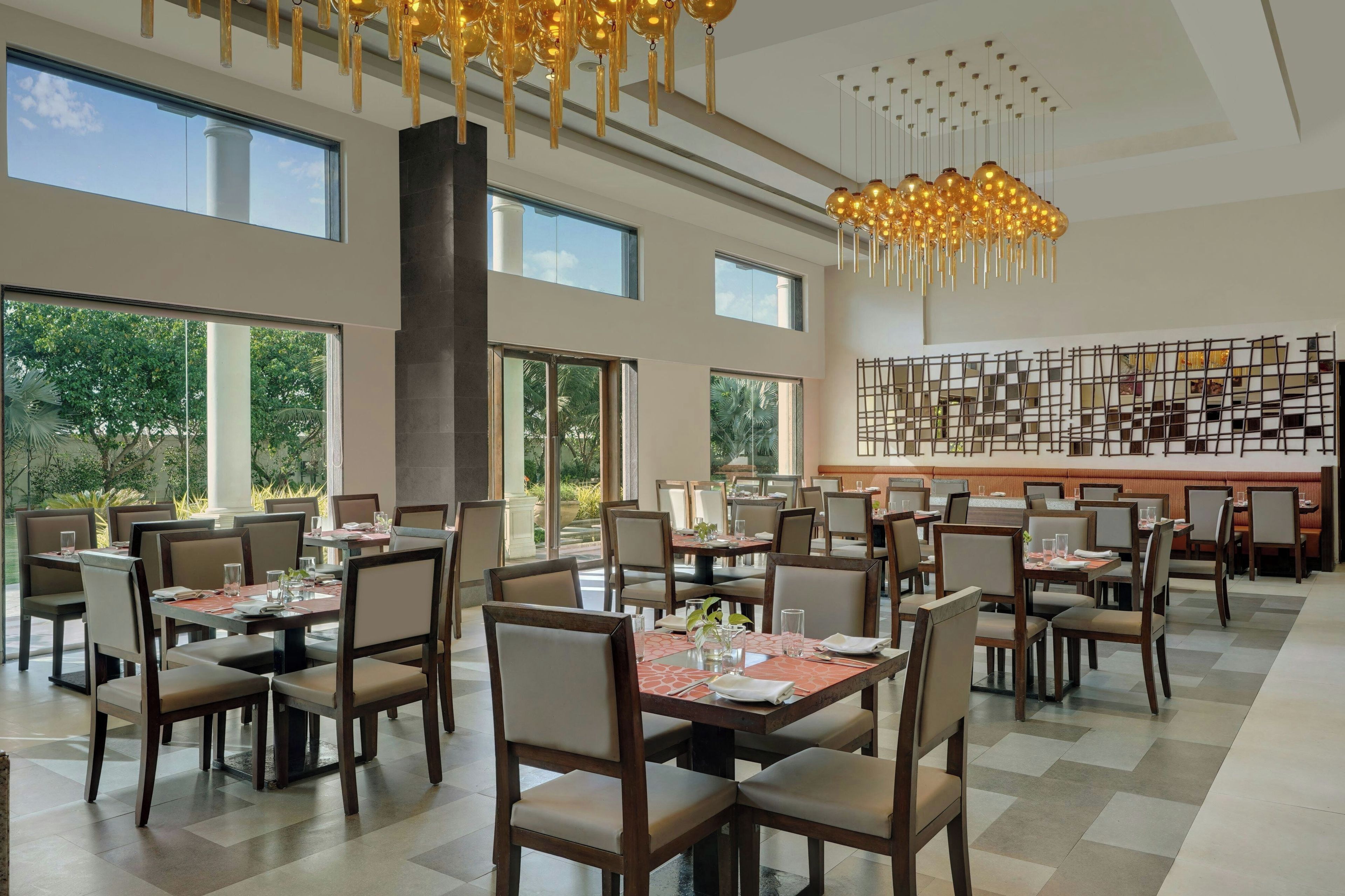 Een ruim restaurant met hoge plafonds, grote ramen en moderne kroonluchters. Tafels zijn netjes gerangschikt met stoelen, en de vloer is betegeld.