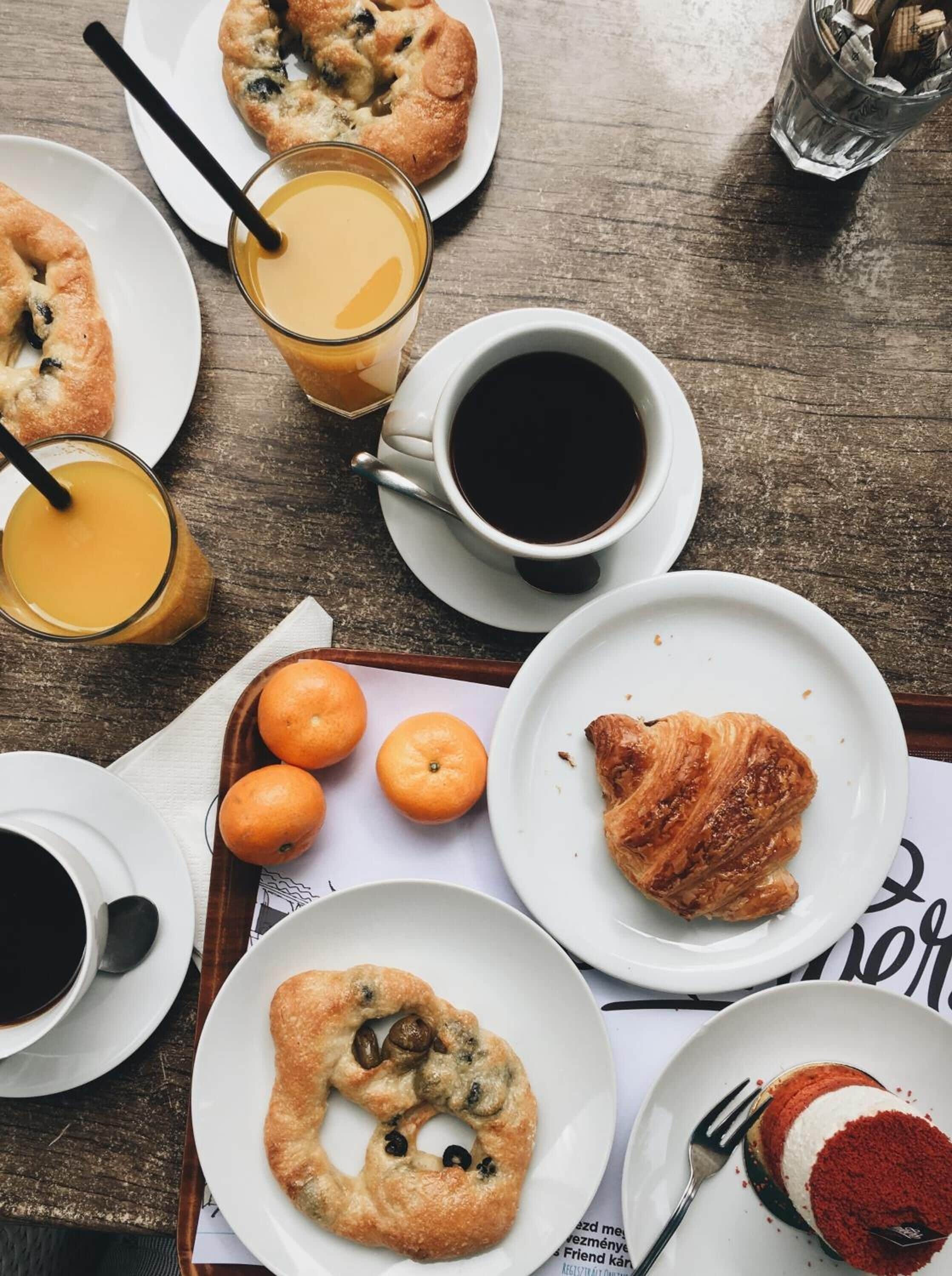 A breakfast setting with croissants, orange juice, coffee, and a dessert on a wooden table.