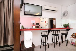 Une cuisine moderne avec un plan de travail rouge, une machine à café, un téléviseur fixé au-dessus du plan de travail et un ventilateur mural.