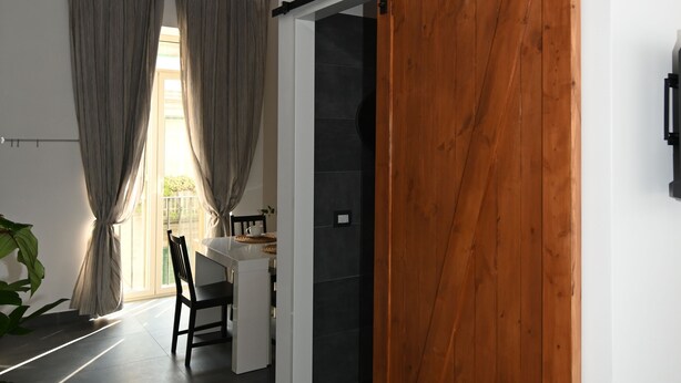 A modern interior with a sliding wooden door, a dining area with a table and chairs, and a view through a window with curtains. - B&B Palummelle (Naples)
