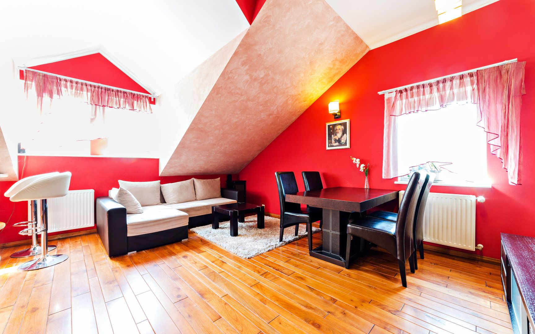 Un appartement moderne de style « loft », avec un plafond en pente, des murs rouges, un plancher en bois, un coin repas avec une table et des chaises, un canapé et un tabouret de bar.