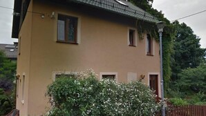 Een huis met twee verdiepingen, een schuin dak, een houten omheining en een struik met witte bloemen ervoor.