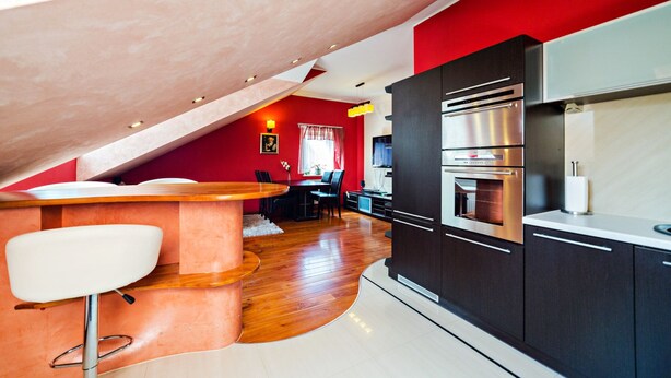 Een moderne keuken met donkere kastjes en roestvrijstalen apparatuur, een houten eettafel met stoelen, en een rode accentmuur.