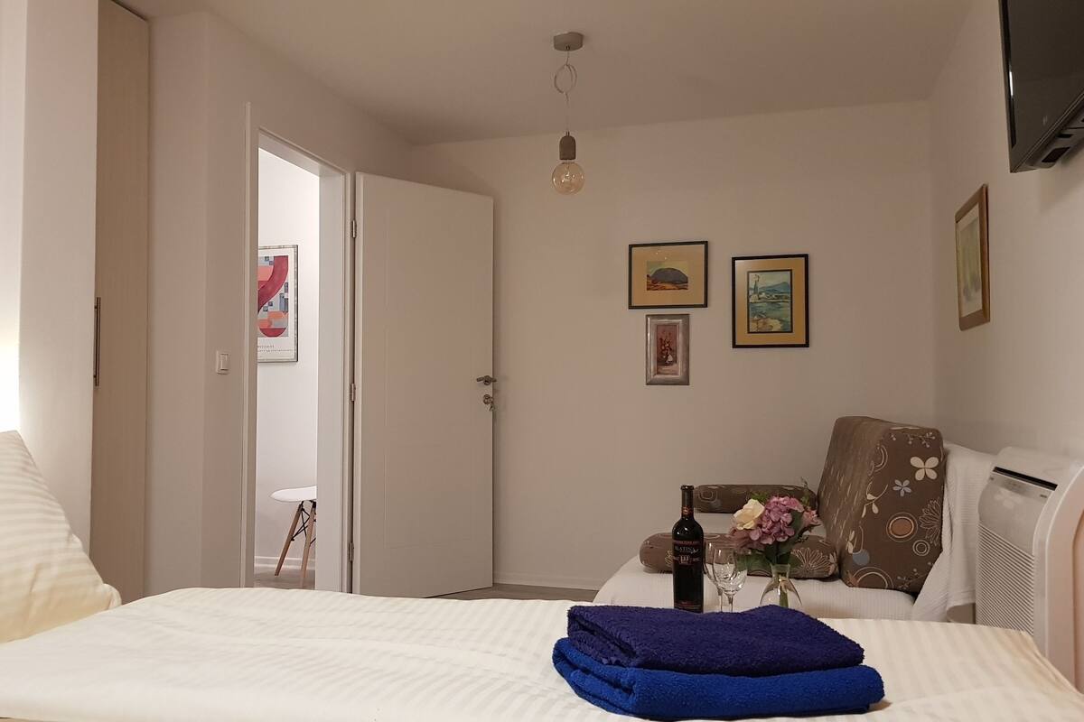 A hotel room with a bed, a chair, a table with a bottle and flowers, and a wall with framed pictures.