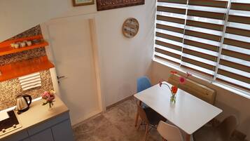 Une cuisine compacte avec un plan de travail blanc, une bouilloire et un vase rempli de fleurs. On y trouve une table à manger avec des chaises et une fenêtre avec des stores.