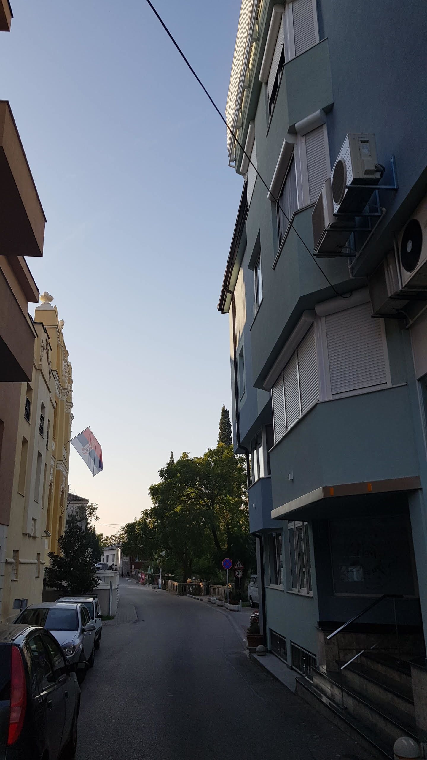 A narrow street with parked cars on one side and a building with air conditioning units and a staircase on the other.