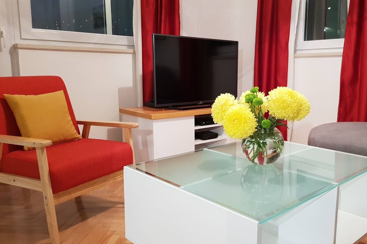 A modern living room with a glass coffee table, a red armchair, a grey sofa, and a TV on a wooden stand.