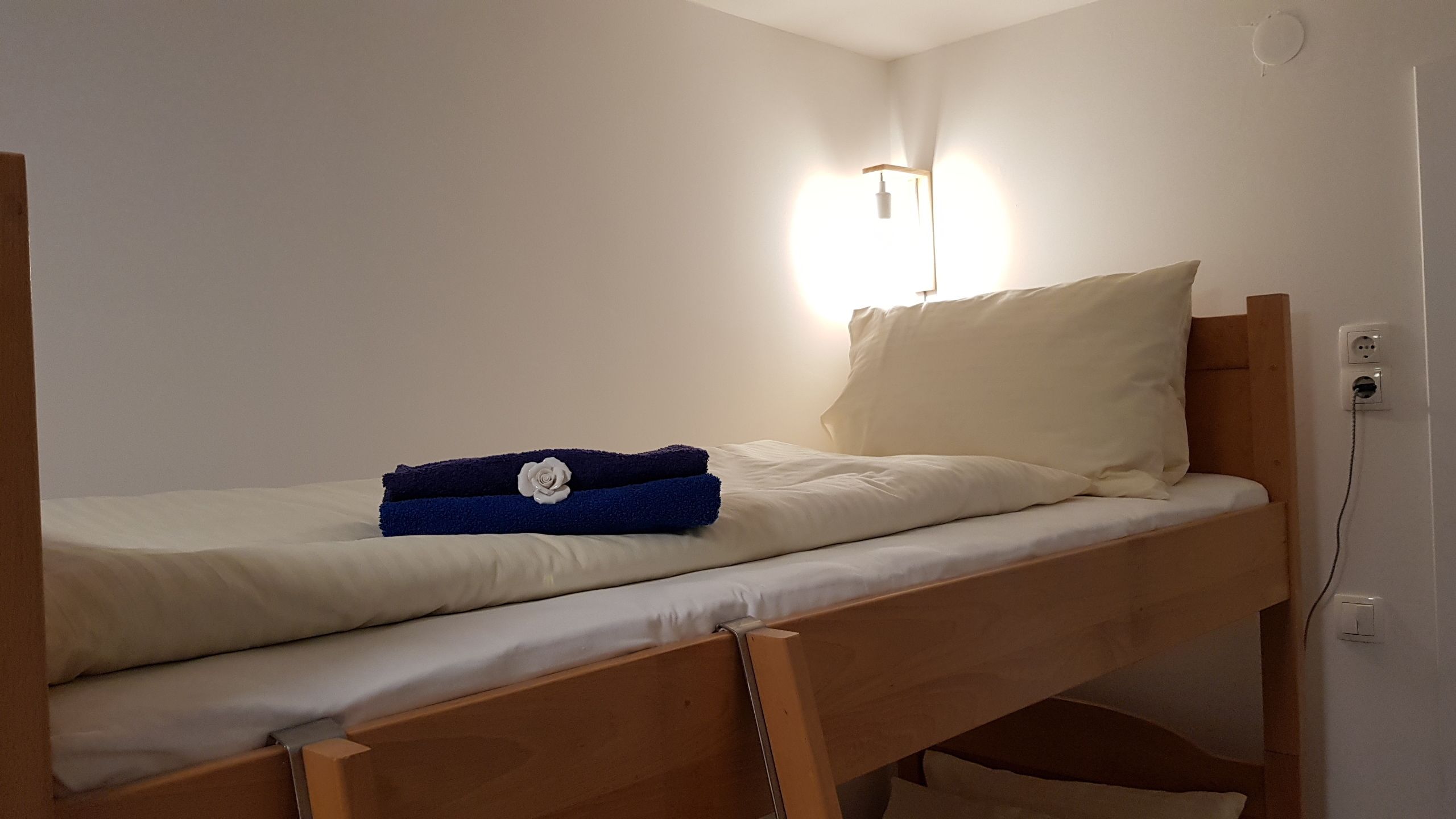 A single bed with a wooden frame, white bedding, and a blue towel with a rose decoration. There is a wall-mounted light above the bed.