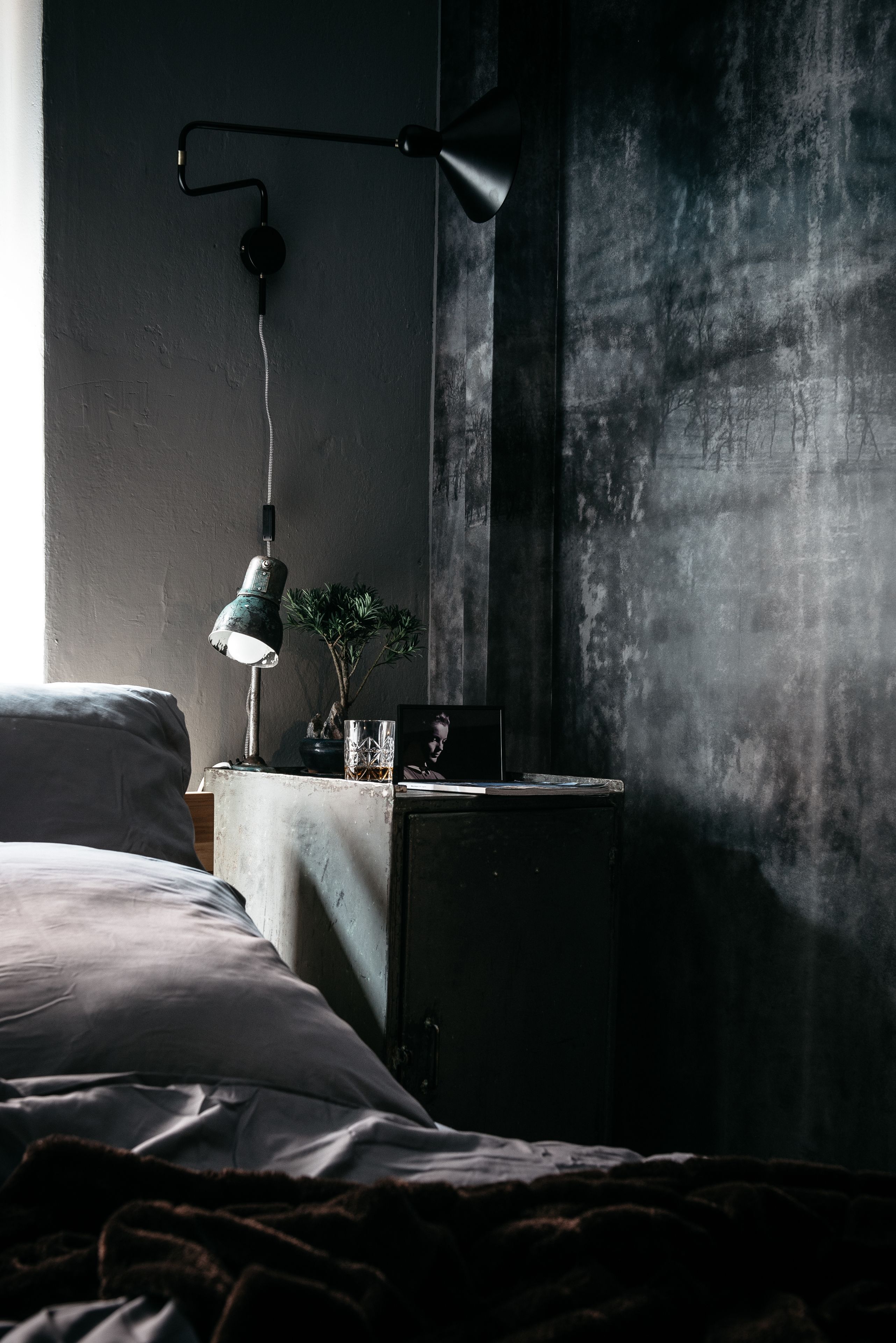 Une chambre à coucher faiblement éclairée, avec un lit, une table de chevet sur laquelle se trouve une lampe, et un luminaire fixé au mur.