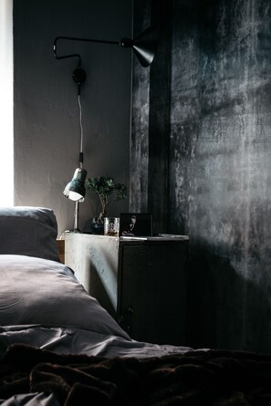 A dimly lit bedroom with a bed, a bedside table with a lamp, and a wall-mounted light fixture. - URBN Dreams (Berlin)