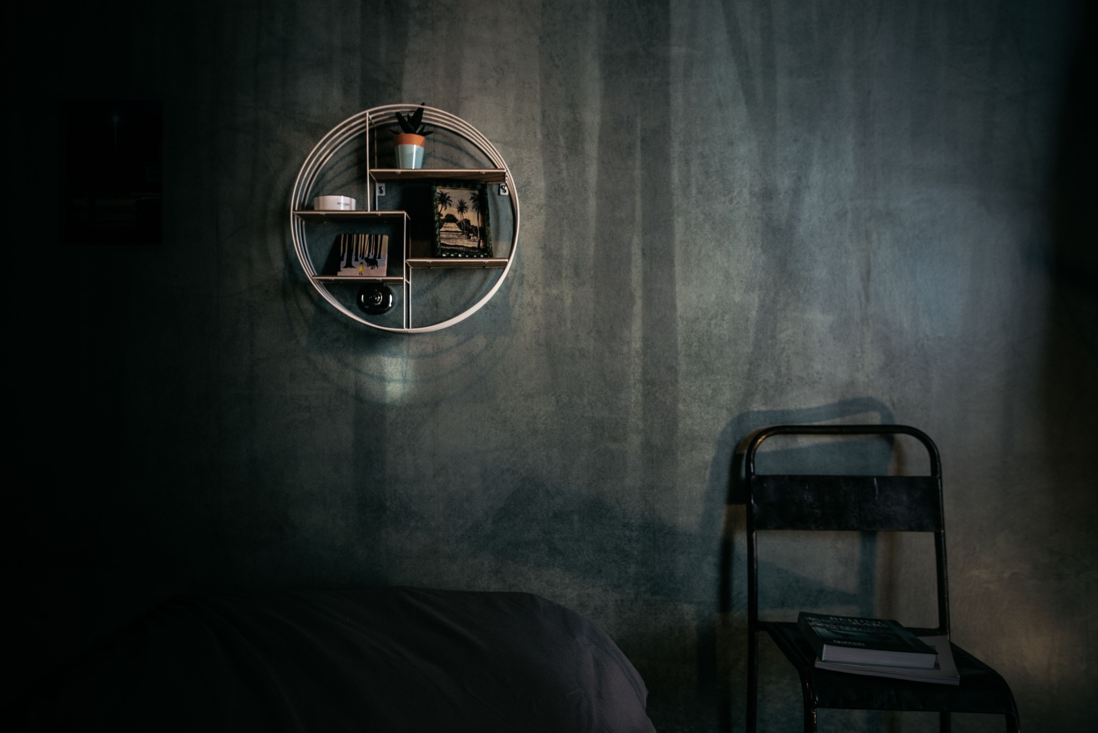 Une étagère circulaire sur laquelle sont disposés divers objets, le tout adossé à un mur à texture, une chaise avec un livre, et un lit dans une pièce faiblement éclairée.