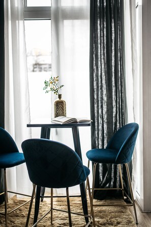 Ein Zimmer mit einem Tisch, zwei blauen Stühlen, einer Vase mit einer Pflanze und einem Fenster mit Vorhängen.
