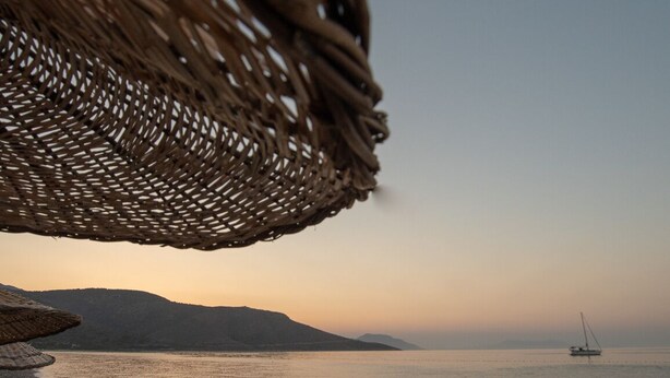 A beach scene with a wicker umbrella, pebbles, and a distant sailboat. - Cuhadar Ahsap Evleri (Datça)