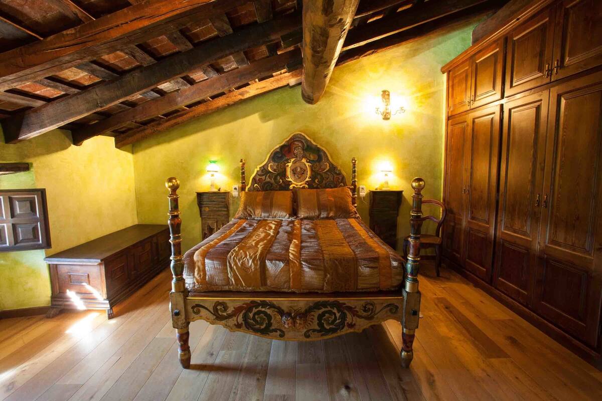Una cama de madera tradicional con intrincados grabados, flanqueada por dos mesitas de noche con lámparas, en una habitación con suelo de madera y techo inclinado.