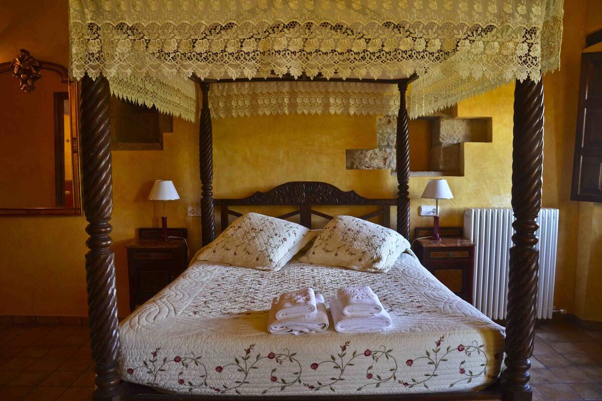 Una cama con dosel adornada con encaje, ropa de cama floral, dos lámparas de noche y un juego de toallas dobladas sobre la cama.