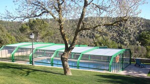 Una hilera de estructuras con techo verde y paredes de vidrio, con una piscina en el centro, rodeada de césped y árboles.