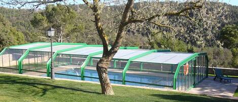 Una hilera de estructuras con techo verde y paredes de vidrio, con una piscina en el centro, rodeada de césped y árboles.