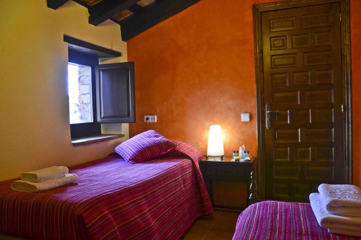 Una habitación con dos camas, cada una con una colcha y almohadas a rayas rojas. Hay una puerta de madera y una ventana con vistas al exterior.
