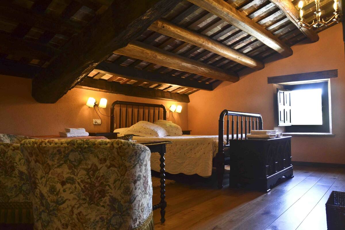 Un dormitorio con techo de madera, una cama con cubrecama blanco, una mesita de noche, una silla y una ventana con luz natural.