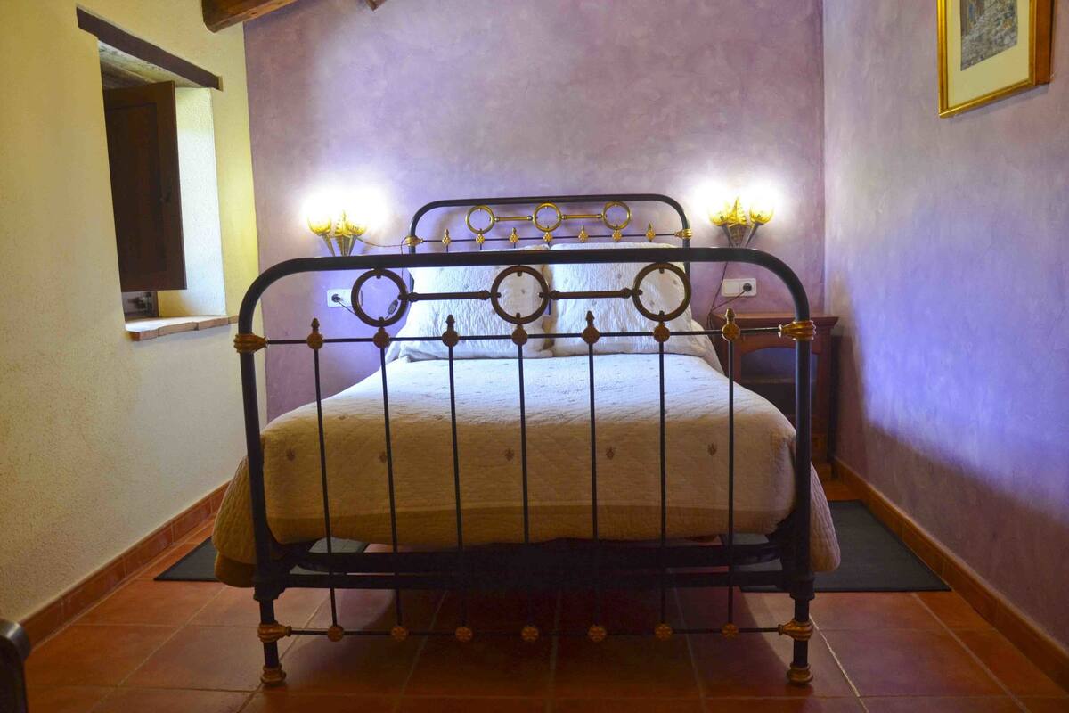 Una cama con estructura metálica y cabecera con diseños circulares, una mesita de noche con lámpara y una ventana a la izquierda.