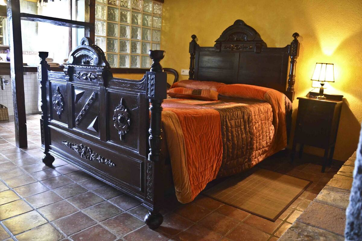 Una cama tradicional con un cabecero oscuro, una mesita de noche con lámpara, y una ventana con vistas al exterior.