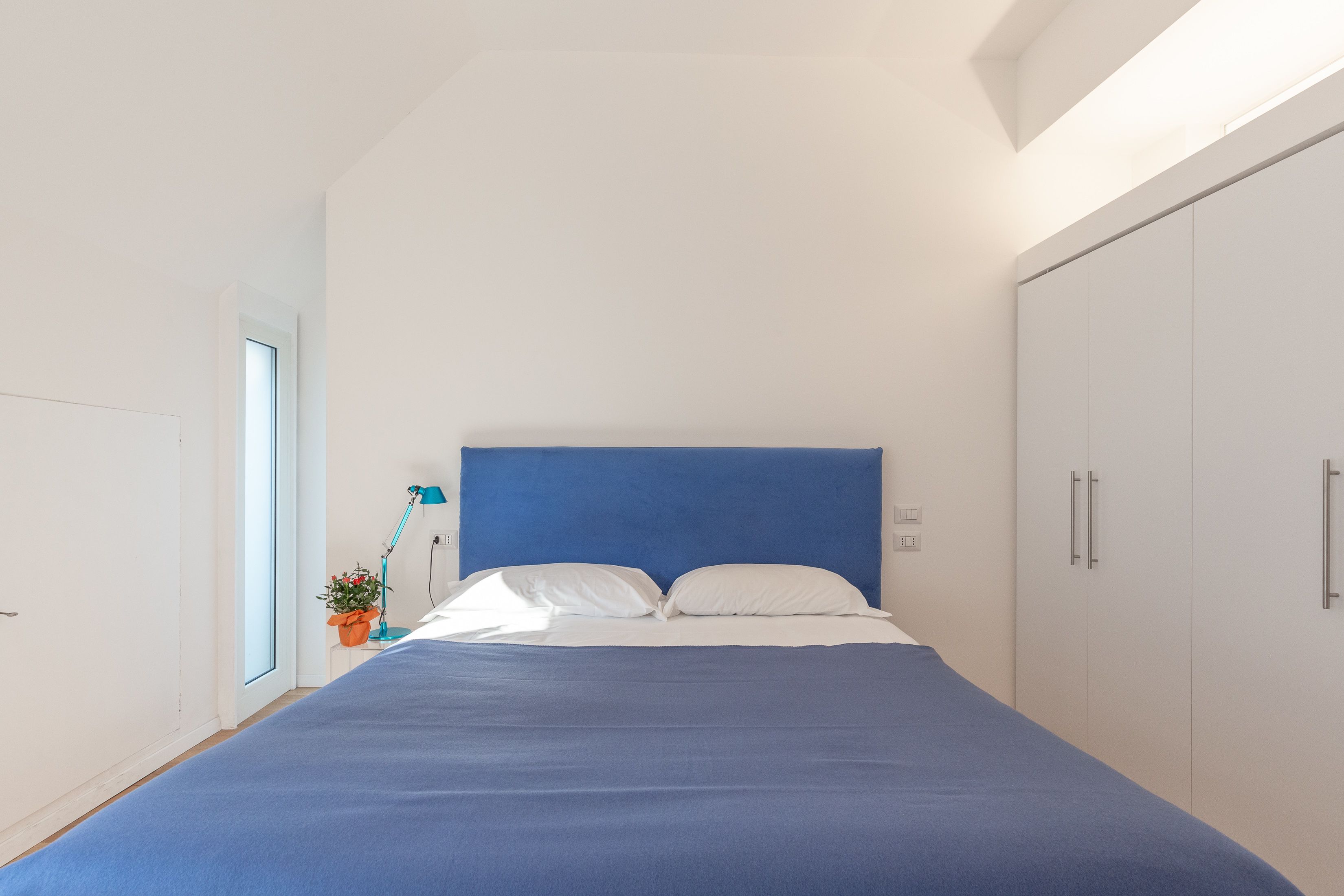 A bedroom with a large bed, a nightstand with a lamp, a potted plant, and a wardrobe.