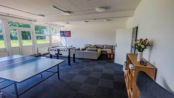 Una habitación con mesa de ping pong, mesa de futbolín, sofá, pequeña mesa con planta y televisor montado en la pared.