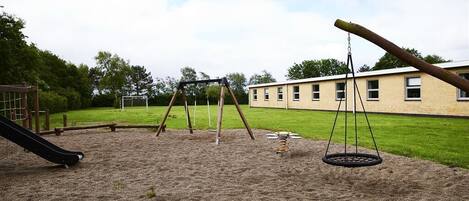 A playground with swings and a slide, set against a building with a grassy field in the background.