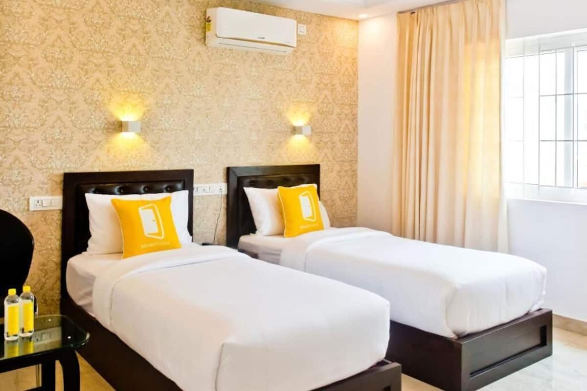 Dos camas individuales con ropa de cama blanca y almohadas amarillas, una mesita con botellas y una ventana con cortinas.