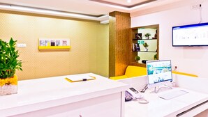 Mostrador de recepción con computadora, teléfono y bloc de notas. Hay una planta a la izquierda y una estantería con libros y otra planta a la derecha.