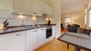 Eine moderne Küche mit weißen Schränken, einer Spüle aus Edelstahl und einem eingebauten Backofen. Ein Essbereich mit Tisch und Stühlen sowie ein Wohnzimmer mit Sofa und Fenster.