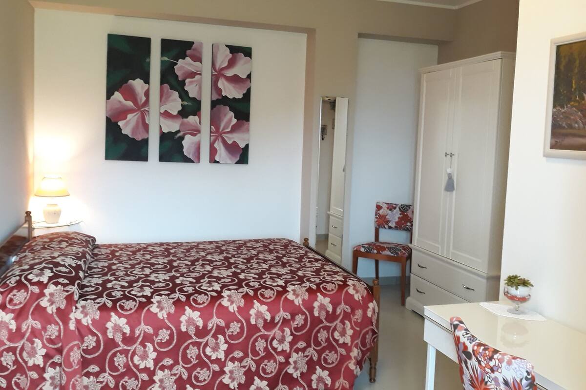 A bedroom with a bed, a desk, a chair, a wardrobe, and a painting on the wall.