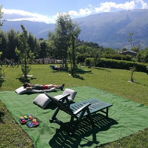 Un espacio al aire libre con césped, sillones de relax y una mesa, una persona acostada sobre un cojín, y pelotas de colores en el suelo.